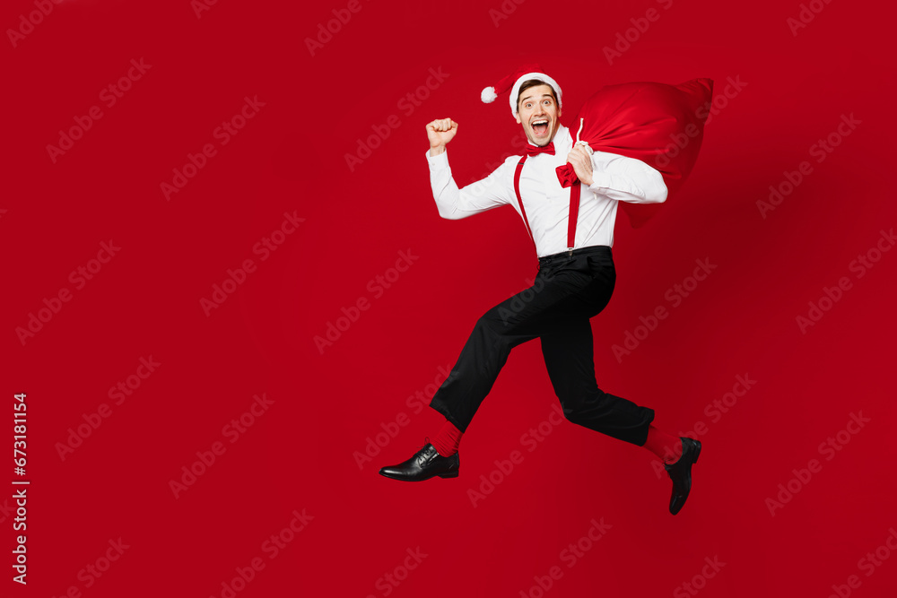 Full body merry young excited man wear white shirt Santa hat posing jump high hold bag with present gifts isolated on plain red background studio. Happy New Year Christmas celebration holiday concept. Full body merry young excited man wear white shirt Santa hat posing jump high hold bag with present gifts isolated on plain red background studio. Happy New Year Christmas celebration holiday concept.