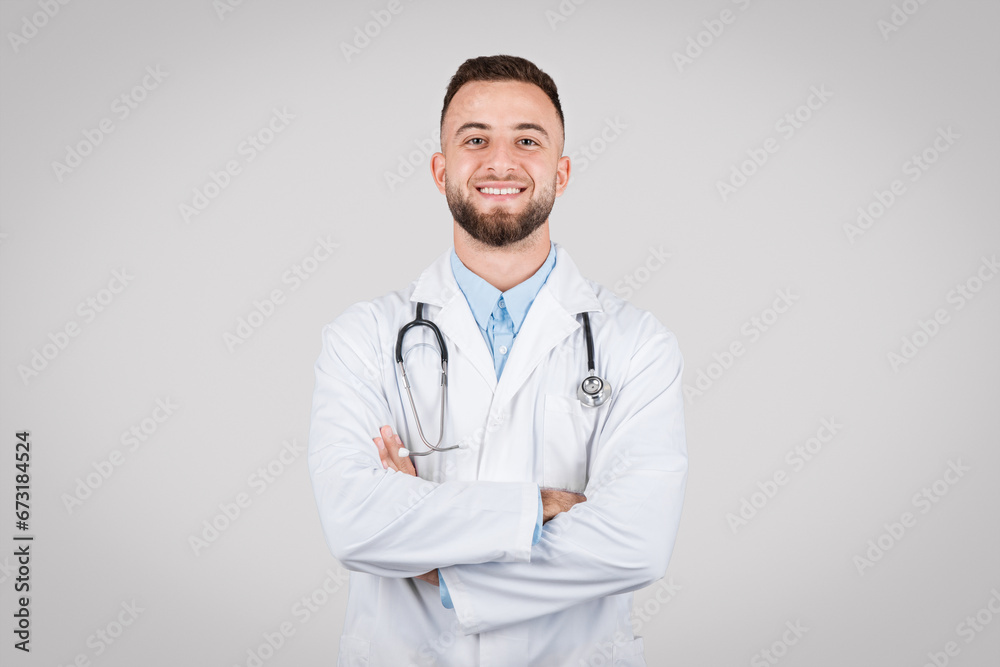 Fototapeta premium Confident young doctor posing with arms crossed and stethoscope around his neck against grey background