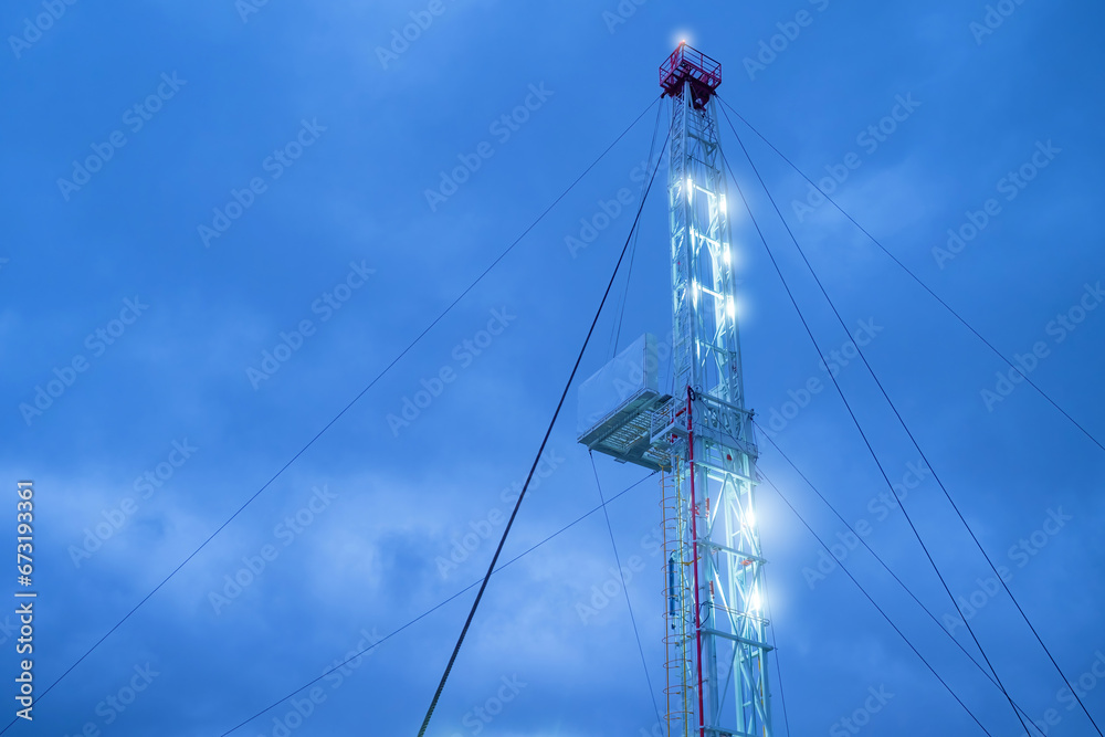 Rig drilling under blue sky. Tower is illuminated by evening lights. Drilling equipment at field. Mining of oil and gas. Petrochemical company drilling rig. Crude oil production. Petroleum industry Rig drilling under blue sky. Tower is illuminated by evening lights. Drilling equipment at field. Mining of oil and gas. Petrochemical company drilling rig. Crude oil production. Petroleum industry