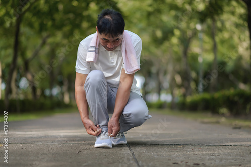 An Elderly Asian Man Is Tying Shoelaces In The Park Full Of Trees Prepare Jogging Or Exercise.