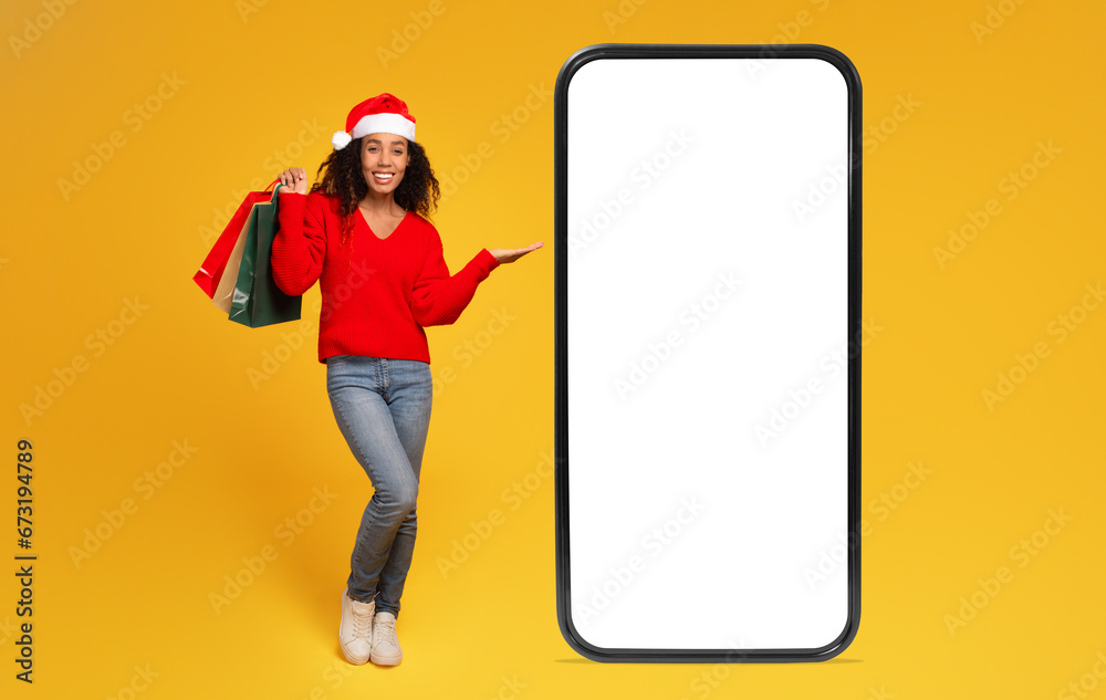 Black lady in Santa hat holds bags and shows phone with blank screen for sales, yellow background - obrazy, fototapety, plakaty 
