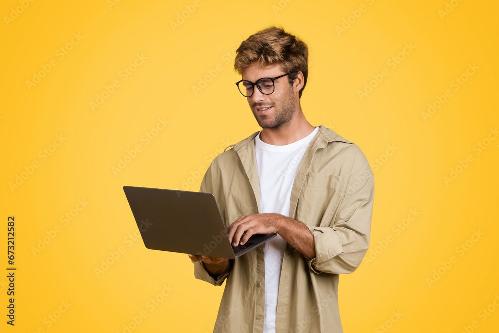 European man typing on laptop, standing against yellow background European man typing on laptop, standing against yellow background