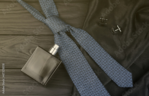 A bottle of mens cologne and cufflinks with blue tie lie on a black luxury fabric background on a wooden table. Mens classic accessories. Shallow DOF
