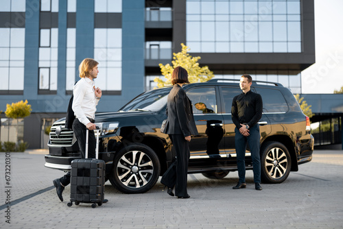 Businessman and businesswoman walk with a suitcase to a luxury black car during a business trip. Male chauffeur waiting near vehicle. Concept of transportation and business travel © rh2010