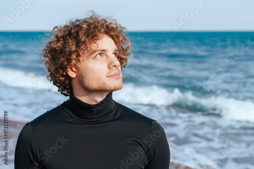 the guy is standing near the sea. a young curly guy in a black turtleneck stands near the blue sea, the guy raised his head towards the sun, a young guy walks along the beach