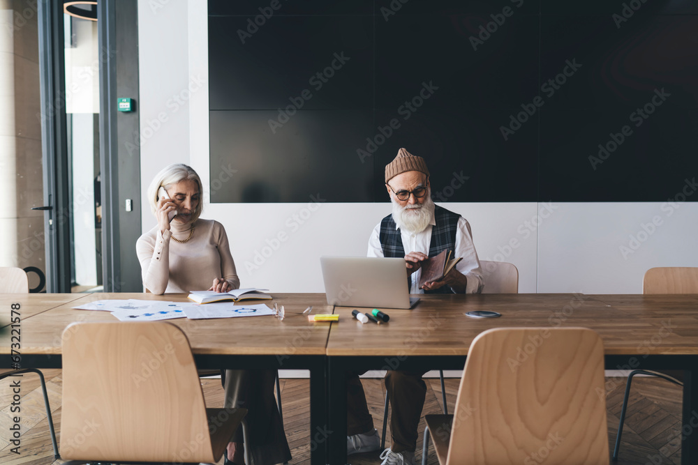 Elderly colleagues sitting at table with diagrams laptop in office Elderly colleagues sitting at table with diagrams laptop in office