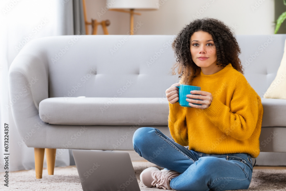 a girl sits on the floor near the sofa with a mug in her hand. a young curly girl in jeans and a yellow sweater sits leaning on the sofa with a blue weight in her hands a girl sits on the floor near the sofa with a mug in her hand. a young curly girl in jeans and a yellow sweater sits leaning on the sofa with a blue weight in her hands