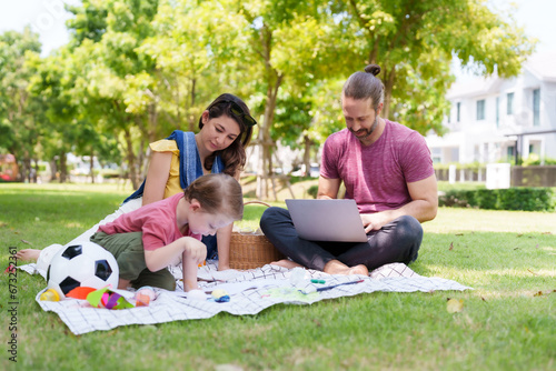 Mother is teaching her child to use a brush to draw and paint. Caucasian family of parents and son is relaxing on grass of village park while the father is working on laptop. family vacation Concept