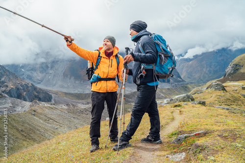 Caucasian and Asian men with backpacks together resting Makalu Barun Park trekking route near Khare. Man pointing at Mera peak summit. Backpackers enjoying climbing acclimatization walk valley view #673256183