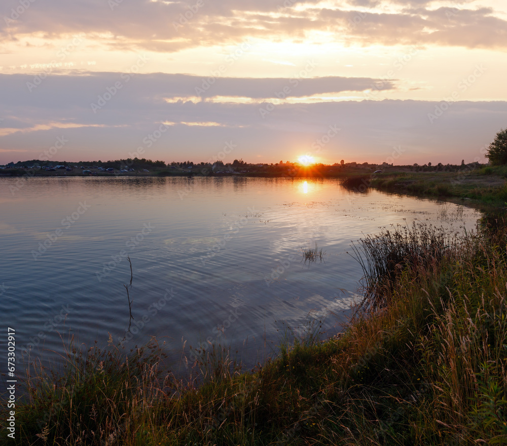 Lake summer sunset view with sun reflection and cars on shore. All people are unrecognizable.
