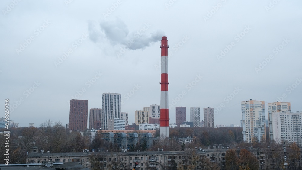 A tall orange and white chimney from which smoke is coming stands in the middle of the city