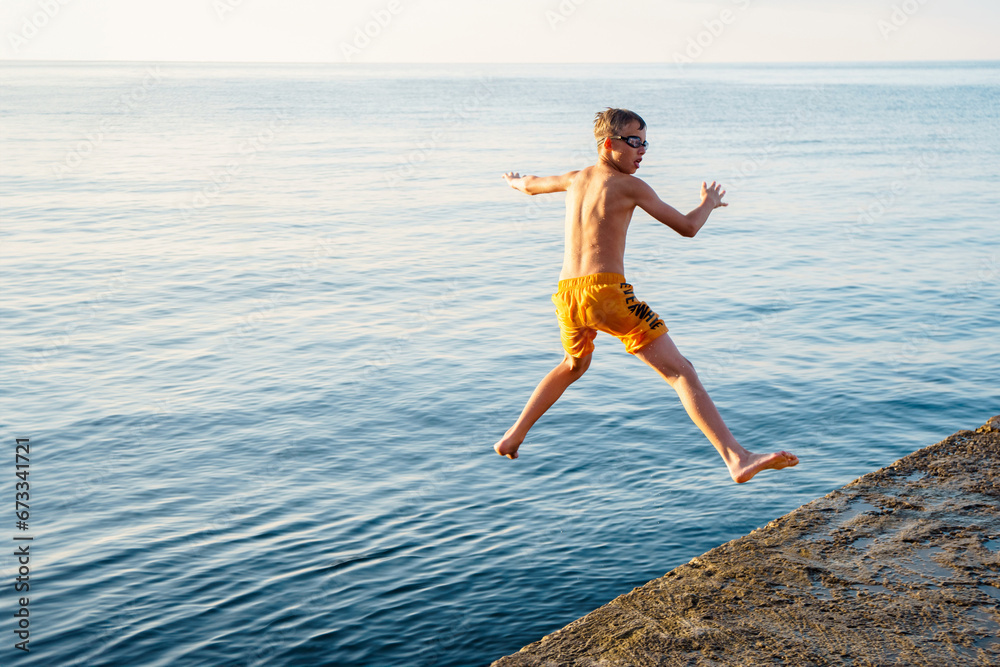 Healthy boy jumps into tranquil ocean water from stone pier in sunny morning. Sportive child dives in sea at summer resort. Active lifestyle