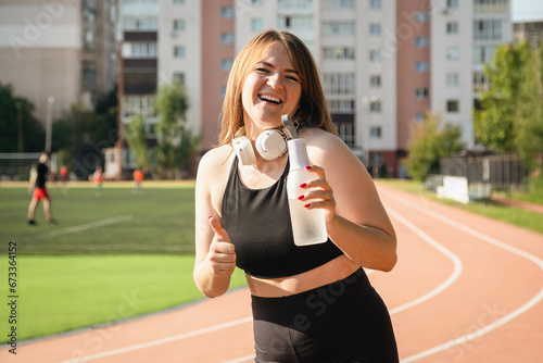 Happy caucasian female athlete stands at the stadium on running track with a bottle of water, training outdoor concept.