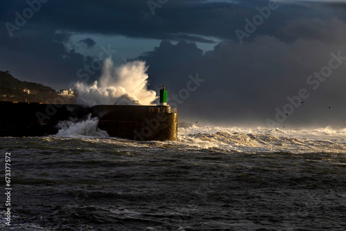 Wave exploding against a lighthouse illuminated by a halo of sunlight about to set. Galicia, Spain.