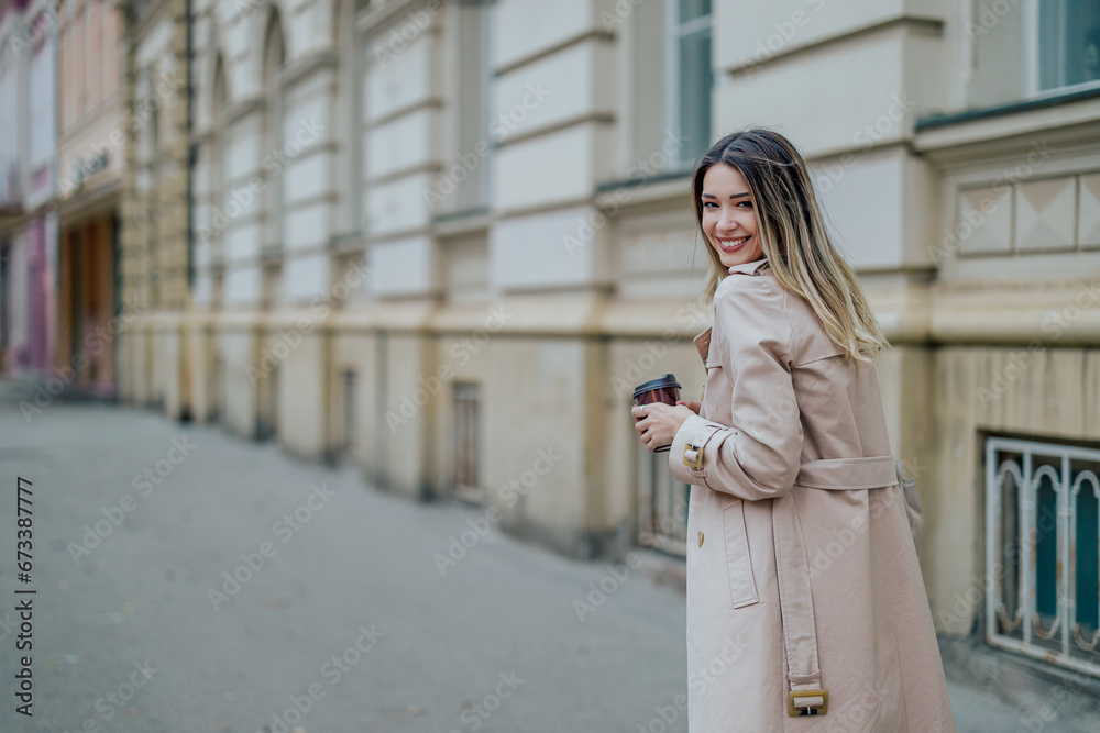 Fototapeta premium Woman in a trench coat walking down the street and drinking coffee takeaway