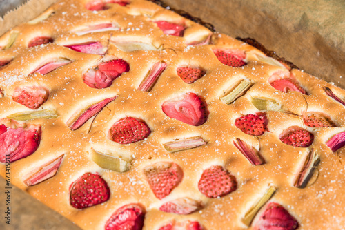 A freshly baked strawberry and rhubarb cake, sprinkled with sugar dusting, captures the essence of summer with its vibrant colors and rich texture.
