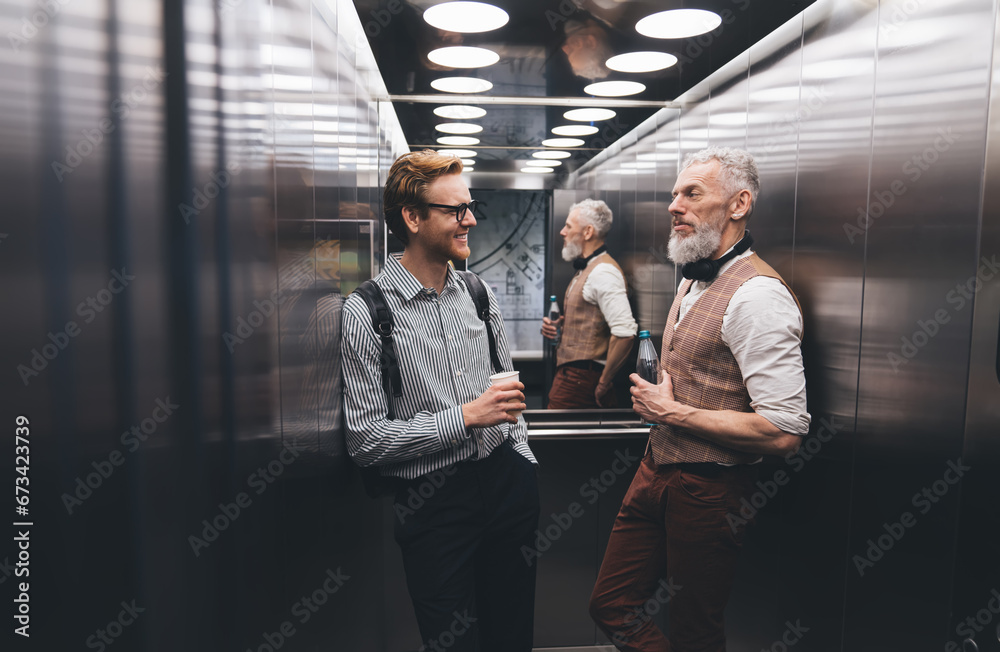 Positive businessman talking with elderly colleague by modern elevator - obrazy, fototapety, plakaty 