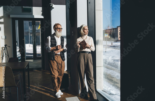 Stylish senior coworkers standing near glass wall in sunlight