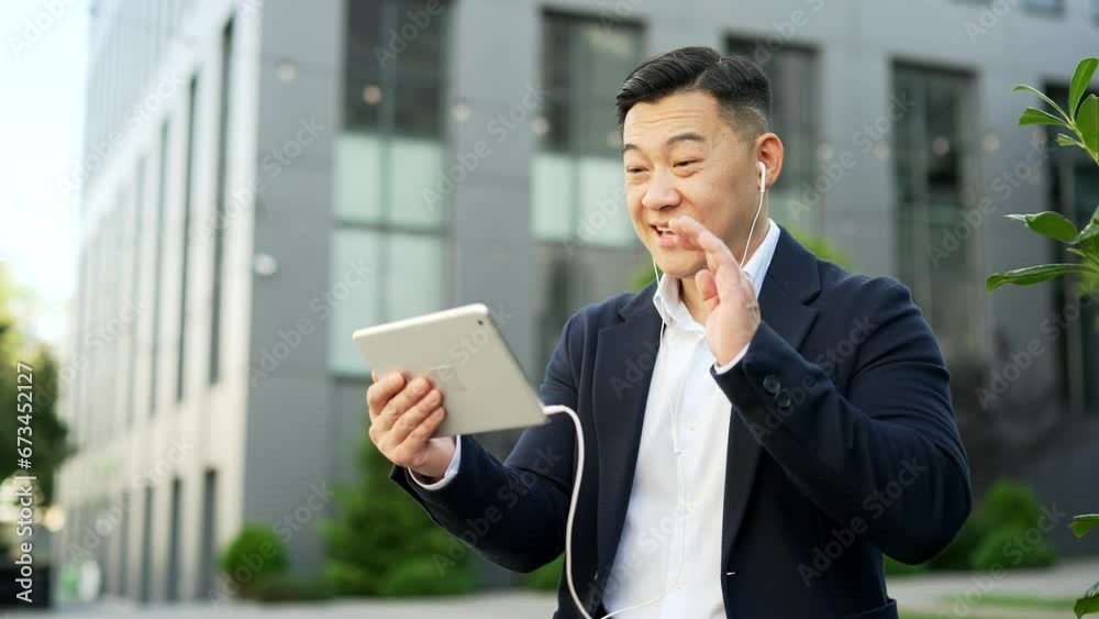 Asian businessman in a formal suit talking on a video call using a digital tablet sitting on a bench on street near office building. Entrepreneur communicates online with a client or business partner Asian businessman in a formal suit talking on a video call using a digital tablet sitting on a bench on street near office building. Entrepreneur communicates online with a client or business partner