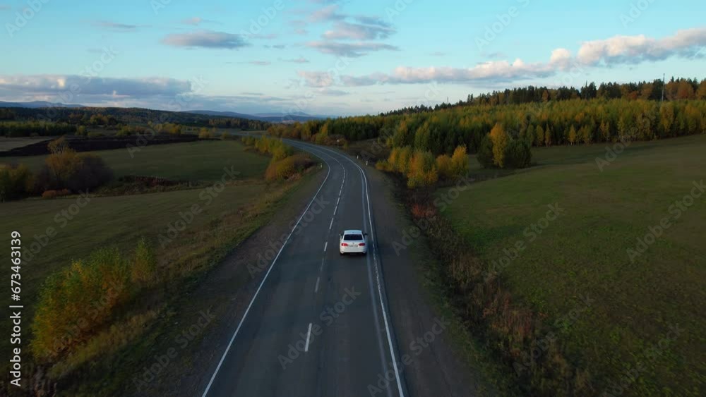One white car driving on an asphalt road in the autumn season, Aerial shot. Picturesque autumn landscapes during sunset. Autumn forest. The drone follows the car One white car driving on an asphalt road in the autumn season, Aerial shot. Picturesque autumn landscapes during sunset. Autumn forest. The drone follows the car