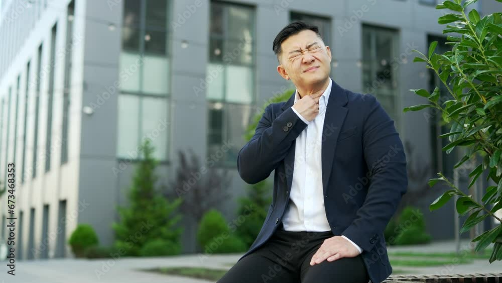 Asian businessman in a formal suit suffering from a sore throat sitting on a bench on the street near an office building. Sick male coughs, feels bad. has a cold, virus or flu, massaging a sore spot Asian businessman in a formal suit suffering from a sore throat sitting on a bench on the street near an office building. Sick male coughs, feels bad. has a cold, virus or flu, massaging a sore spot