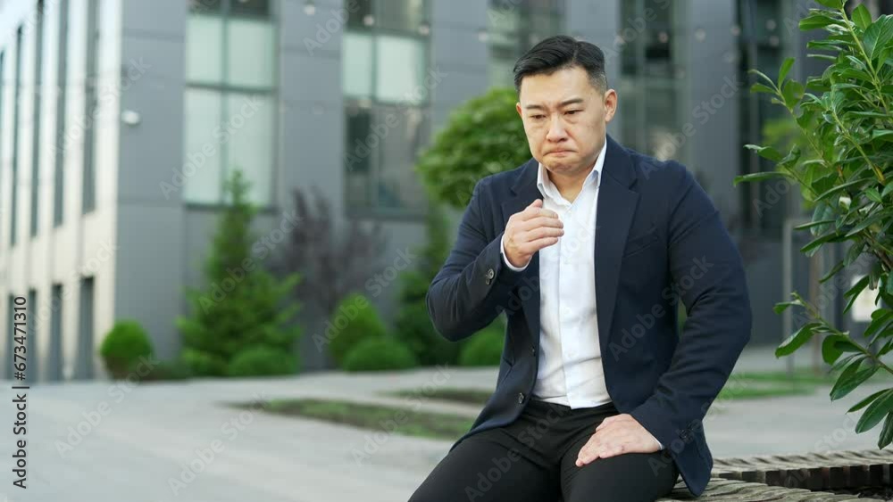 Asian businessman in formal suit suffering from nausea, wants to vomit while sitting on a bench on the street near an office building. Unhealthy male feels bad, has a stomach ache. He has poisoning Asian businessman in formal suit suffering from nausea, wants to vomit while sitting on a bench on the street near an office building. Unhealthy male feels bad, has a stomach ache. He has poisoning