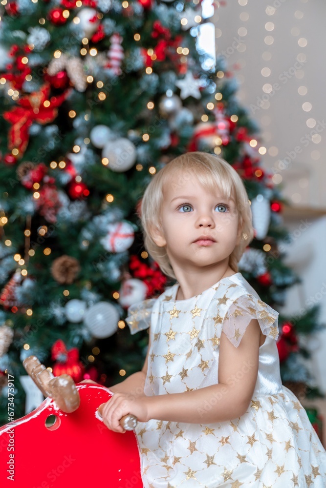 little girl Christmas tree. Blonde 2 years old in a dress sits on a wooden red horse in a room decorated for christmas and new year. Photo under the tree. little girl Christmas tree. Blonde 2 years old in a dress sits on a wooden red horse in a room decorated for christmas and new year. Photo under the tree.