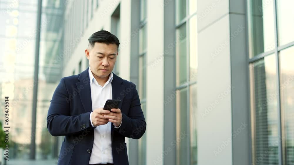 Confident businessman in formal suit is using a smartphone while walking on the street near an office building. Entrepreneur is reading email messages, chatting online, texting with a business partner