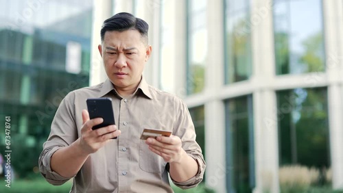 Shocked asian businessman checks his credit card on smartphone standing on street near office building. Frustrated male faced fraud, money was stolen from his account. He became a victim of deception photo
