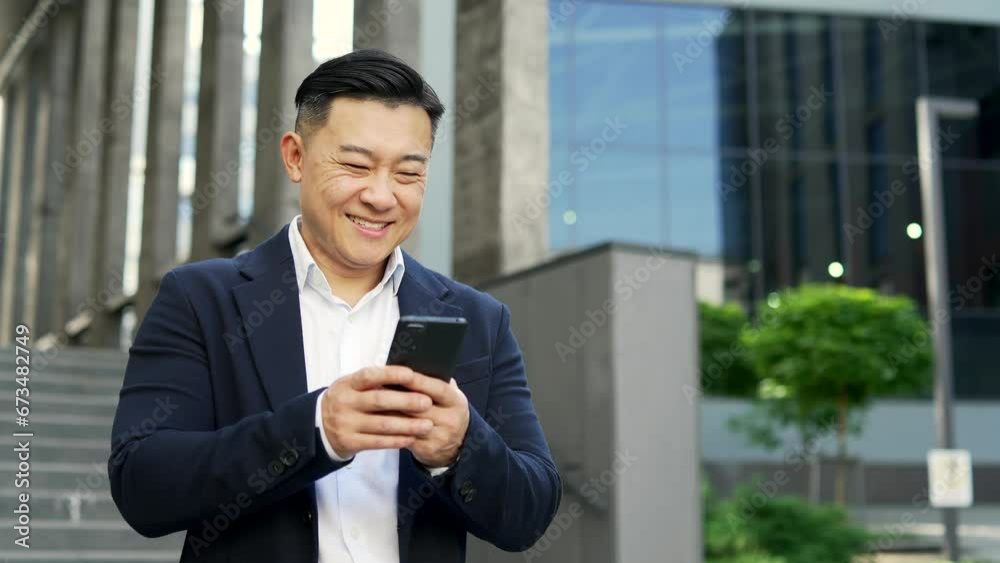 Smiling businessman in formal suit is using a smartphone while standing on the street near an office building. Happy entrepreneur browsing the phone, checking email messages, chatting online, texting Smiling businessman in formal suit is using a smartphone while standing on the street near an office building. Happy entrepreneur browsing the phone, checking email messages, chatting online, texting