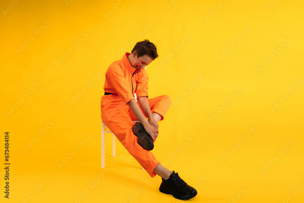 Middle age funny and Cheerful woman wear modern Orange overalls sitting on white chair over yellow color background. Female having fun and energetic movement studio shot Middle age funny and Cheerful woman wear modern Orange overalls sitting on white chair over yellow color background. Female having fun and energetic movement studio shot