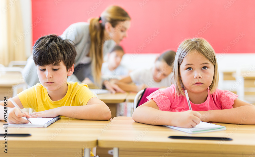 Upset serious preteen boy and girl listening to teacher and writing exercises in notebook at lesson in primary school
