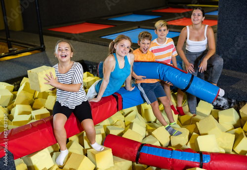Company of joyful smiling children sitting together on crossbar in entertainment trampoline centre surrounded with foam cubes © JackF