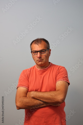 A self-assured middle-aged man, sporting eyeglasses and crossing his arms, is dressed in an orange t-shirt against a plain gray background.