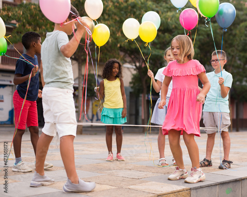 Multiracial group of cheerful preteen children having fun together outdoors on summer day, playing chinese jump rope with colorful toy balloons in hands photo