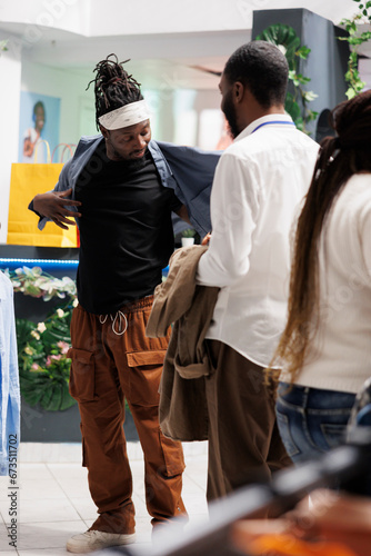 Shopping mall assistant helping customer to try clothes before buying in fashion showroom. Young african american man wearing shirt while checking apparel size in modern store