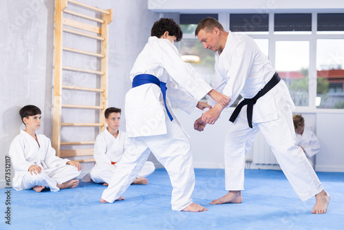 Experienced male karate instructor demonstrating attacking techniques in sparring with young martial arts student to group of teenagers sitting on tatami during training..