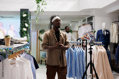 African american influencer filming live video on smartphone to showcase clothes in store, speaking to followers on camera. Blogger promoting apparel collection for social media photo