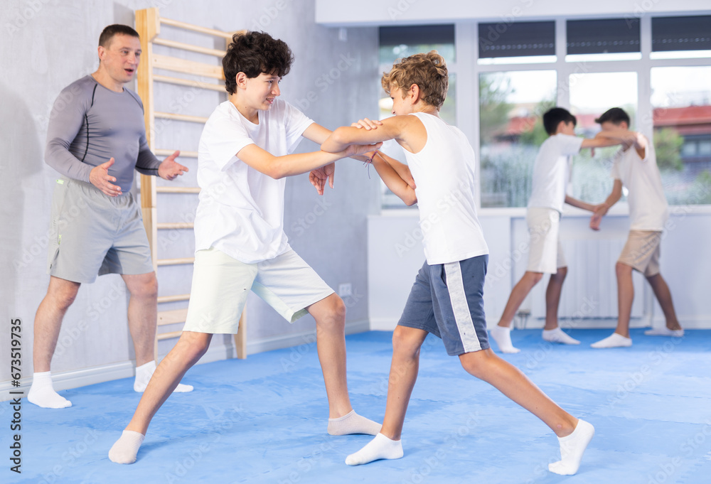 Focused teenage boys practicing close combat techniques in sparring in training room during self-defence workout under supervision of coach