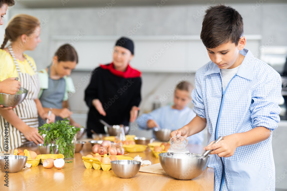 During culinary classes, teen boy combine essential components in mixing bowl and readies dough for bread rolls. In background, blurry children stand near kitchen table and listen to chef explanations
