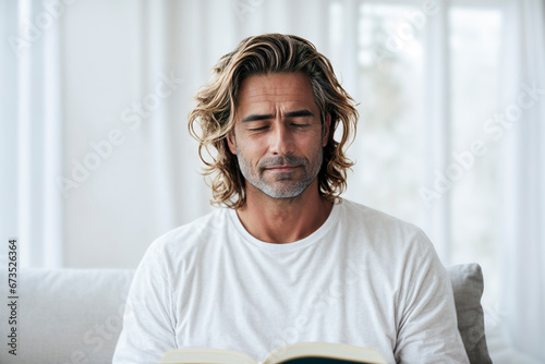 A man with shoulder-length hair and a white T-shirt reads a book, eyes closed, looking peaceful.