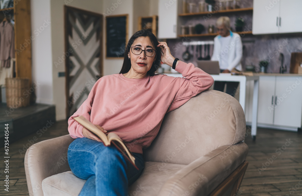 Obraz premium Cheerful middle aged ethnic woman sitting with book on sofa
