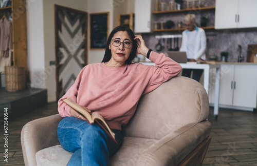Cheerful middle aged ethnic woman sitting with book on sofa