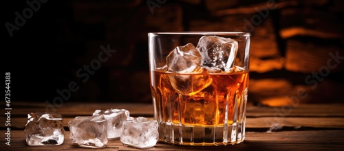 A beverage containing 2 ounces of scotch or bourbon poured over ice cubes commonly known as on the rocks is a shot of amber spirits with an ice cube resting on a wooden countertop alongside  photo