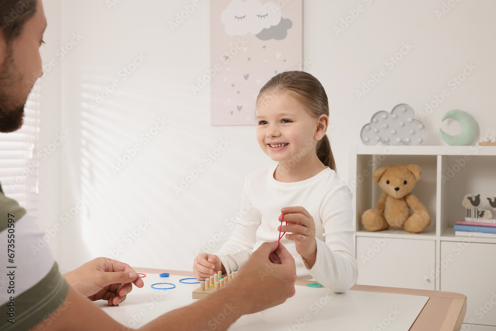 Motor skills development. Father helping his daughter to play with geoboard and rubber bands at white table in room Motor skills development. Father helping his daughter to play with geoboard and rubber bands at white table in room