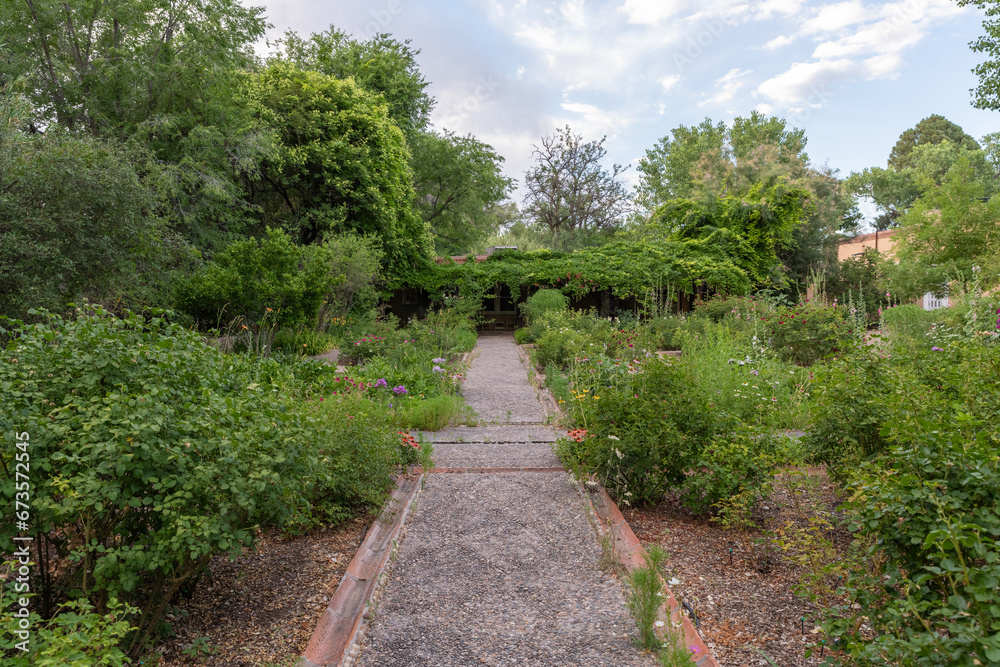 A gravel walking path lined with bushes and wildflowers leads to an ivy covered entryway in a tranquil garden, with green trees and a partly cloudy sky in the background. A gravel walking path lined with bushes and wildflowers leads to an ivy covered entryway in a tranquil garden, with green trees and a partly cloudy sky in the background.