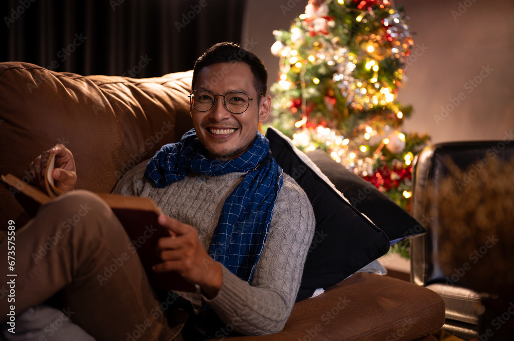 A happy man is celebrating his Christmas night alone, reading a book on a couch in his living room. A happy man is celebrating his Christmas night alone, reading a book on a couch in his living room.