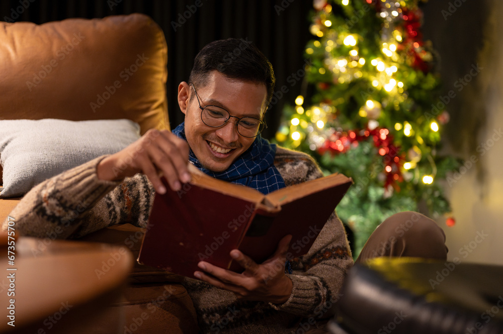 A cheerful Asian man is enjoying reading a book in his living room on Christmas night.