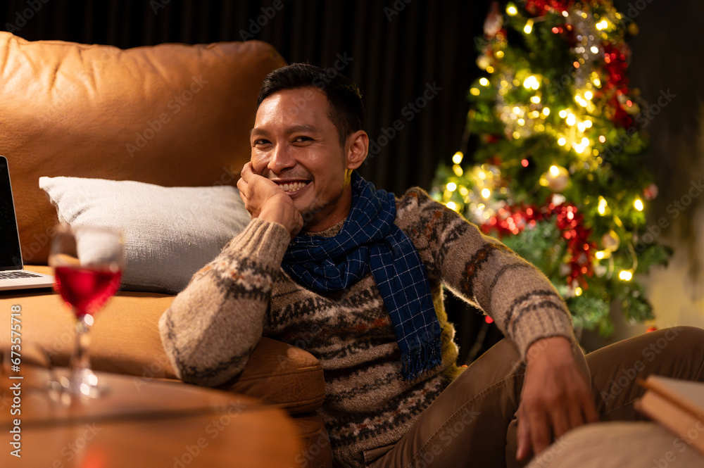 Happy Asian man in comfy winter clothes sitting beside a sofa in his living room on Christmas night