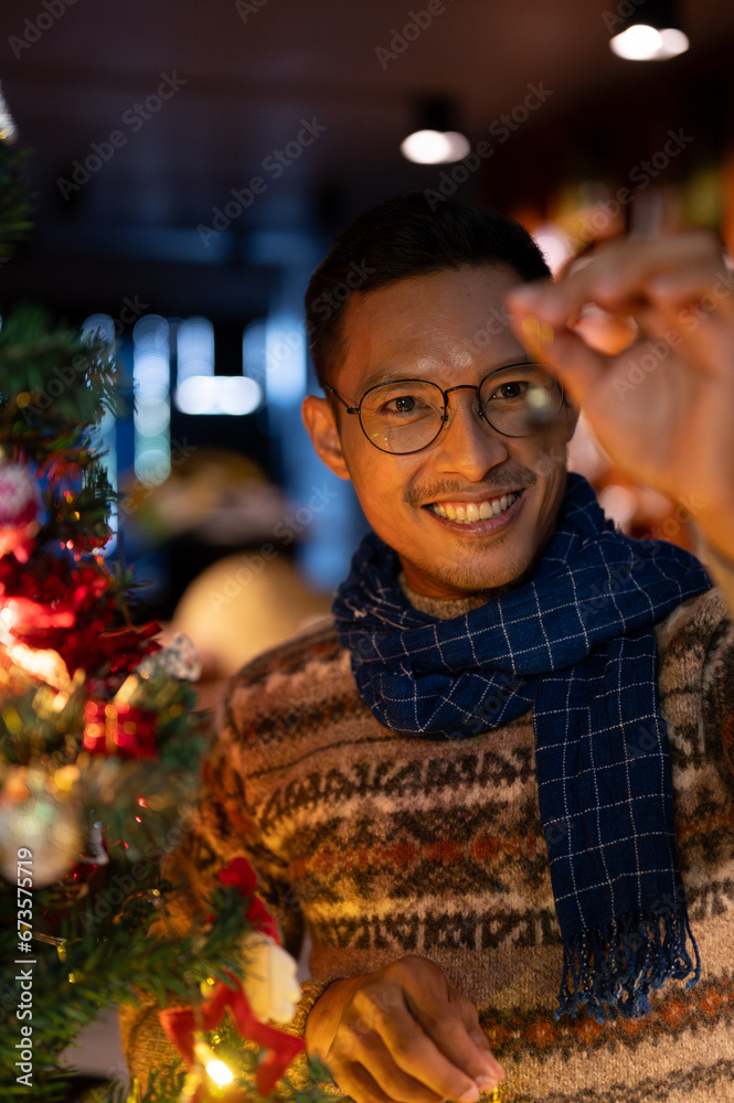 A handsome Asian man is admiring a beautiful Christmas ornament, decorating his Christmas tree.